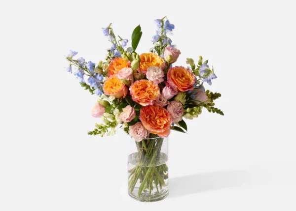 Bouquet of orange and pink roses with blue delphiniums in a clear glass vase on a white background