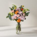 Bouquet of pink, orange, and yellow flowers in a clear glass vase on a light table surface.