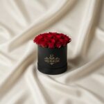 Black round box overflowing with red roses sits on cream satin fabric, labeled 'Les Fleurs'.