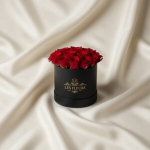 Black round box overflowing with red roses sits on cream satin fabric, labeled 'Les Fleurs'.