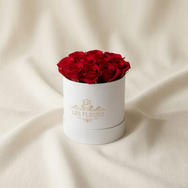 Red rose bouquet arranged in a white round hatbox on pale fabric background.