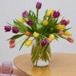 Glass vase of mixed yellow, purple, and pink tulips in water on a round wooden table.