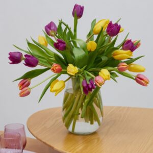 Glass vase of mixed yellow, purple, and pink tulips in water on a round wooden table.