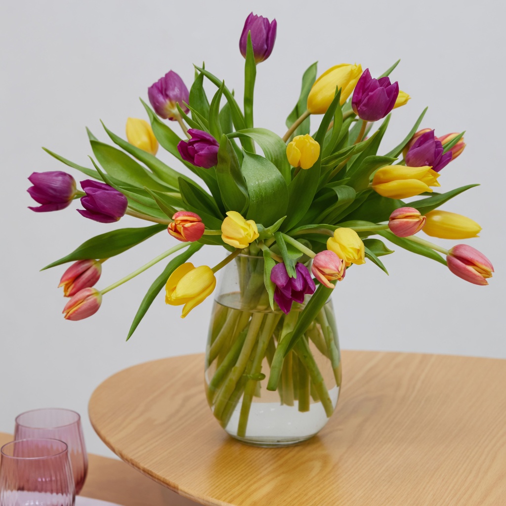 Glass vase of mixed yellow, purple, and pink tulips in water on a round wooden table.