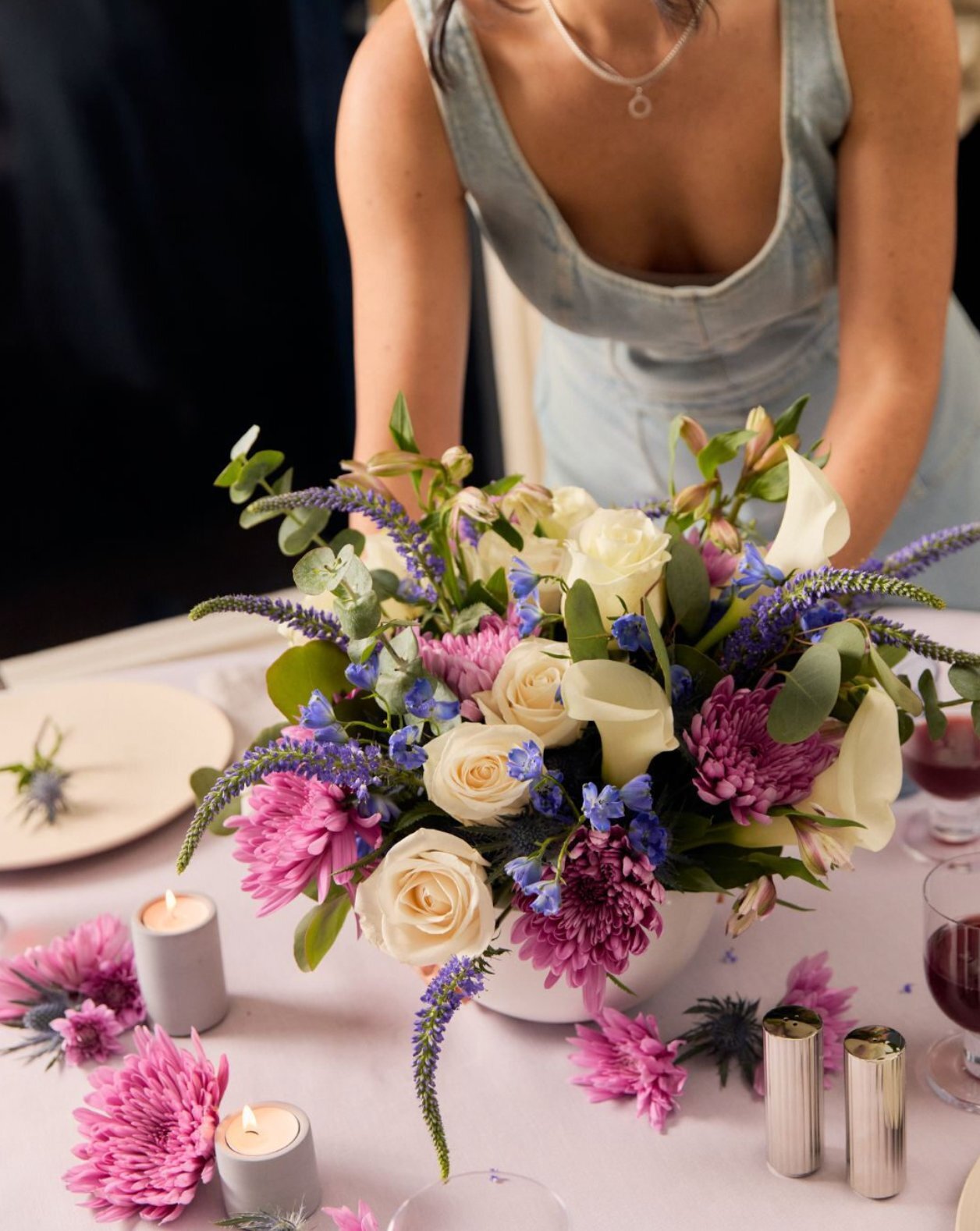 Mujer colocando centro floral con rosas blancas, crisantemos morados y delphinium azul en mesa con velas para Día de la Madre