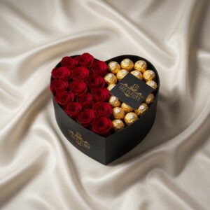 Heart-shaped black gift box with red roses on one half and gold-wrapped chocolates on the other, cream satin backdrop and a 'Les Fleurs' card folded on top.